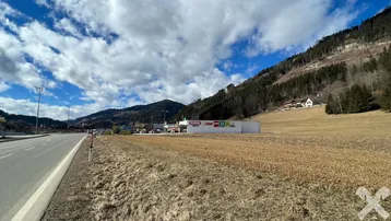 Weitläufige Landschaft mit einem Feld, einer Straße und einem Geschäftshaus vor einer bewaldeten Bergkulisse unter bewölktem Himmel.