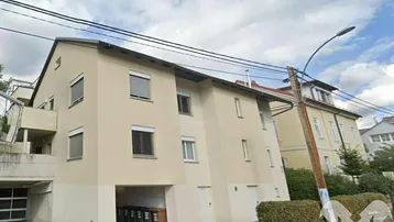 Mehrfamilienhaus mit beiger Fassade, Rollläden und Tiefgaragenzufahrt unter bewölktem Himmel.