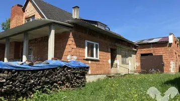 Unfertiges Backsteingebäude mit Vordach und Holzstapel im Vordergrund, umgeben von einer Wiese.