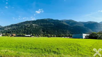 Weitläufige grüne Wiesenfläche mit bewaldeten Bergen im Hintergrund und einigen Gebäuden in der Ferne.