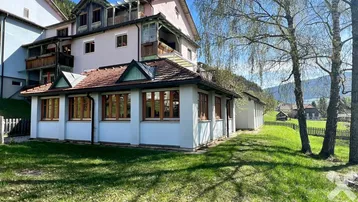 Gebäude mit weißer Fassade und rosa Obergeschoss, umgeben von grüner Wiese und Bäumen mit Bergblick.