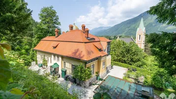 Panoramablick auf die historische Villa mit rotem Ziegeldach und die umliegende Berglandschaft mit Kirche.
