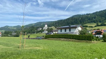 Weitläufige grüne Landschaft mit einem modernen Haus, umgeben von Hügeln und einem Dorf im Hintergrund.