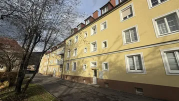 Gelbe Fassade eines Mehrfamilienhauses mit vielen Fenstern und Mansarddach unter bewölktem Himmel.