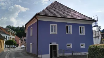 Blau gestrichenes Gebäude mit Satteldach und Rampe zum Eingangsbereich an einer Straße.