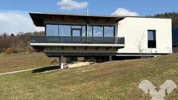 Modernes Haus mit großen Glasfronten und Balkon auf einem Hanggrundstück mit weitem Blick.