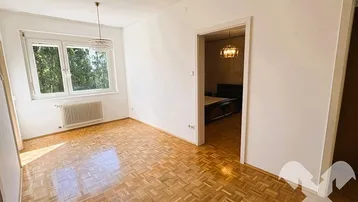 Heller Raum mit Parkettboden und Fenster, der zu einem angrenzenden Schlafzimmer führt.