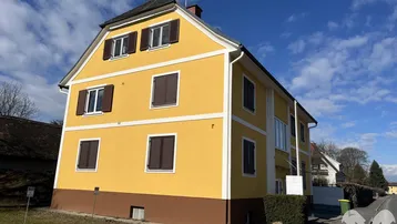 Gelbes Gebäude mit braunem Sockel und dunklen Fensterläden unter blauem Himmel.