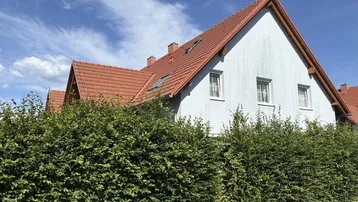 Reihenhausfassade mit hellblauer Farbe und rotem Ziegeldach, umgeben von einer grünen Hecke unter blauem Himmel.