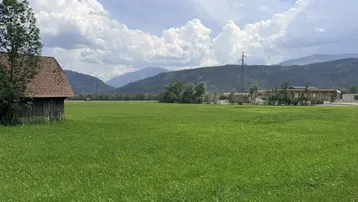 Weitläufige grüne Wiese mit einem kleinen Holzschuppen und Bergen im Hintergrund unter bewölktem Himmel.