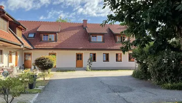 Traditionelles Gebäude mit rotem Ziegeldach und gepflasterter Zufahrt, umgeben von Grünflächen.
