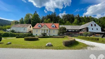 Gelbes Landhaus mit rotem Dach, umgeben von einer gepflegten Rasenfläche und weiteren Gebäuden in ländlicher Umgebung.