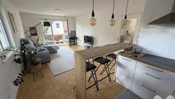 Heller offener Wohnbereich mit integrierter Küche, Parkettboden und Zugang zum Balkon.