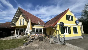 Gelbes Gebäude mit rotem Ziegeldach und gepflastertem Vorplatz unter blauem Himmel.