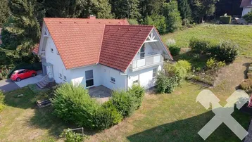 Weißes Einfamilienhaus mit rotem Ziegeldach, Balkon und großem Garten in grüner Umgebung.