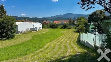 Große grüne Wiese mit gemähten Bahnen, umgeben von Gebäuden und Bergen im Hintergrund unter blauem Himmel.
