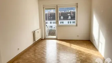 Helles Zimmer mit Parkettboden und Zugang zu einem kleinen Balkon, ideal als Wohn- oder Schlafzimmer.