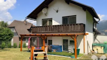 Haus mit Garten, überdachter Terrasse und Balkon, umgeben von grüner Landschaft.
