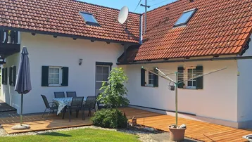 Helle Holzterrasse mit Gartenmöbeln und Sonnenschirm, angrenzend an das Haus mit roten Ziegeldächern.