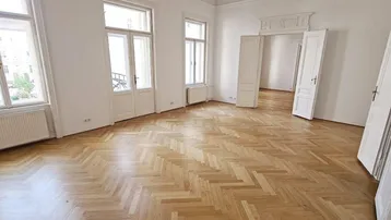 Helles Zimmer mit Fischgrätparkett, hohen Decken und großen Fenstern in einem Altbau.