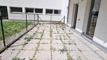 Ungepflegte Terrasse mit Betonplatten und Unkraut, umgeben von einem Zaun und Rasen.