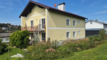 Gelbes Einfamilienhaus mit Balkon, Wendeltreppe und gepflegtem Gartenbereich unter blauem Himmel.
