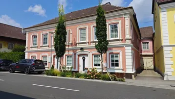 Historisches Gebäude mit rosa Fassade, weißen Fensterrahmen und zwei hohen Bäumen davor.