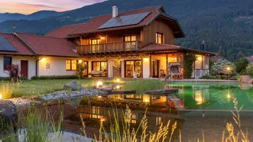 Haus mit beleuchtetem Garten, Naturpool und Bergblick bei Sonnenuntergang.