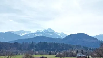 Panoramablick auf schneebedeckte Berge und grüne Hügel mit vereinzelten Häusern in der Ferne.
