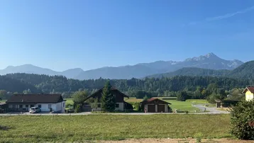 Malerische ländliche Szenerie mit mehreren Häusern, weiten Feldern und einer beeindruckenden Bergkette am Horizont.