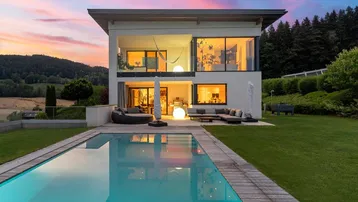 Moderne Villa mit großem Pool und Terrasse, umgeben von grüner Landschaft bei Sonnenuntergang.