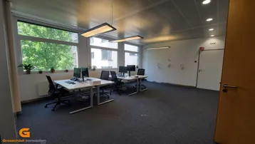 Helles Büro mit mehreren Arbeitsplätzen, großen Fenstern und Teppichboden, ideal für ein Team.