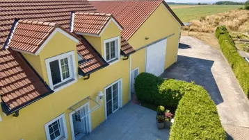 Luftaufnahme des gelben Hauses mit rotem Ziegeldach, gepflasterter Terrasse und angrenzendem Feld.