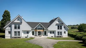 Großes, modernes Einfamilienhaus mit weißer Fassade und dunklem Dach, umgeben von Grünflächen.