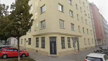 Gebäude an einer Straßenecke mit hellgelber Fassade und vielen Fenstern, umgeben von Bäumen und parkenden Autos.