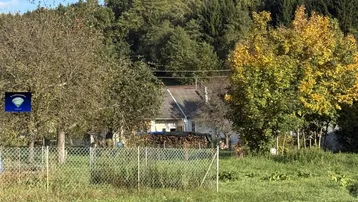 Blick auf ein ländliches Haus mit Holzlager, umgeben von Bäumen und Wiesen unter blauem Himmel.