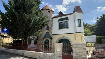 Historisches Gebäude mit markantem Steinturm, weißer Fassade und grünen Fensterläden unter blauem Himmel.