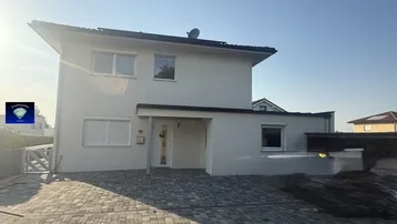 Modernes Einfamilienhaus mit heller Fassade, Rollläden und gepflasterter Einfahrt unter blauem Himmel.