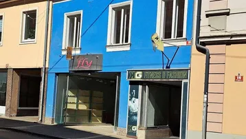 Blau gestrichene Fassade eines Gebäudes mit großen Fenstern und Ladenflächen im Erdgeschoss.