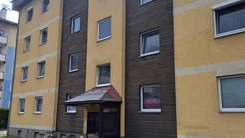 Mehrstöckiges Wohngebäude mit gelber Fassade und dunklen Holzverkleidungen unter bewölktem Himmel.