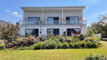Moderne Hausfassade mit mehreren Balkonen, einem gepflegten Garten und blühenden Sträuchern unter blauem Himmel.