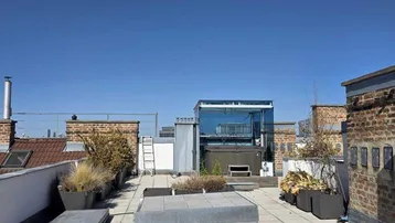 Große Dachterrasse mit Steinplatten, Pflanzenkübeln und einem verglasten Whirlpool unter blauem Himmel.