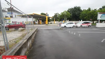 Außenansicht eines Parkplatzes neben einer Tankstelle mit umliegenden Gebäuden und Bäumen.