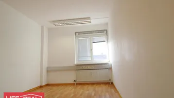 Heller, leerer Raum mit Holzfußboden, Fenster und Heizkörper, ideal für ein Büro.