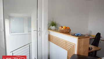 Moderner Bürobereich mit hellem Holzschreibtisch, Laptop und Blick auf ein Treppenhaus durch eine Glastür.
