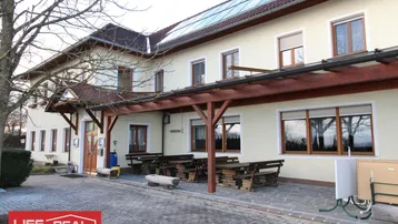 Gebäudeaußenansicht mit gepflastertem Vorplatz, Holztischen und Bänken sowie Solarpaneelen auf dem Dach.