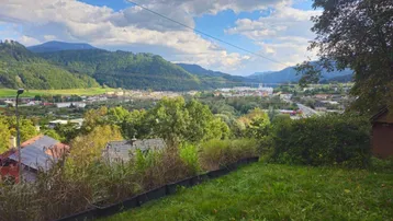 Panoramablick über das Tal mit einer Siedlung und umliegenden grünen Bergen unter bewölktem Himmel.