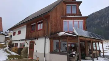 Traditionelles Holzhaus mit weißer Fassade, roten Fenstern und Solarpaneelen auf dem Dach.