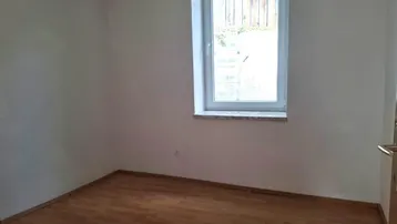 Ruhiges Zimmer mit Laminatboden und Fensterblick auf eine Holzwand und Grünflächen.