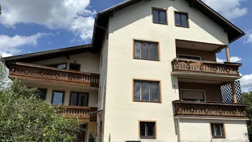 Wohnhaus mit mehreren Balkonen und traditionellen Holzdetails, umgeben von üppigem Grün.